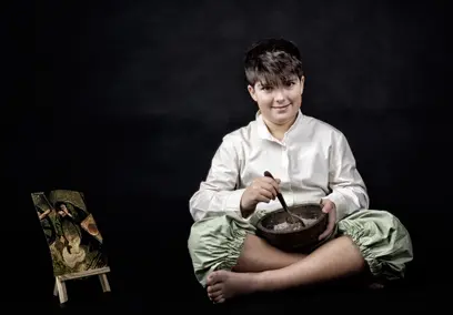 Flemish proverb photo: boy in a light shirt sitting cross-legged on a black background, stirring a wooden bowl of porridge