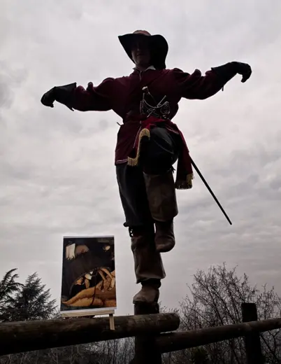 Flemish proverb photo: man in musketeer costume balancing on one leg on a wooden fence, arms outstretched, cloudy sky