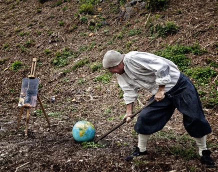 Flemish proverb photo: man in period costume hits a globe with a hoe next to an easel with a Bruegel painting detail