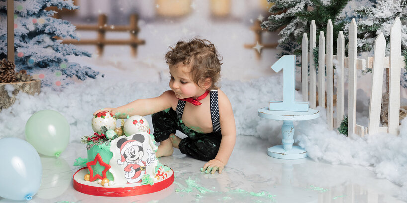 Bimbo festeggia il suo primo compleanno con una sessione fotografica smash cake nel mio studio. Indossa pantaloni bretelle e un papillon rosso mentre si diverte con una torta di Topolino, sporcandosi di panna verde