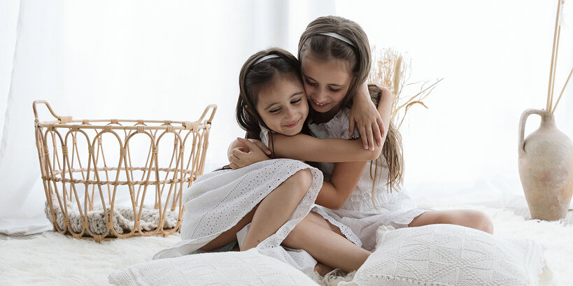 Due dolci sorelline si abbracciano teneramente su un tappeto in uno studio fotografico in luce naturale, indossando adorabili vestitini chiari in un ambiente shabby chic.
