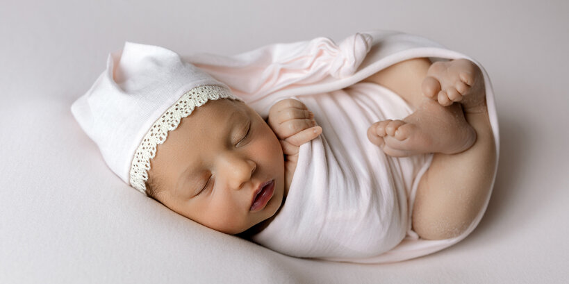 Bambina appena nata dorme serenamente fasciata in un drappo rosa e con un cappellino da notte durante una sessione fotografica newborn nel mio studio
