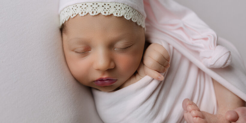 Bambina appena nata dorme serenamente fasciata in un drappo rosa e con un cappellino da notte durante una sessione fotografica newborn nel mio studio