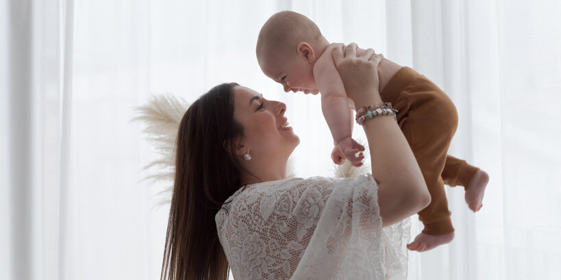 Mamma felice alza il suo bimbo piccolo con gioia, con sfondo di tende bianche nell'atmosfera shabby chic dello studio fotografico