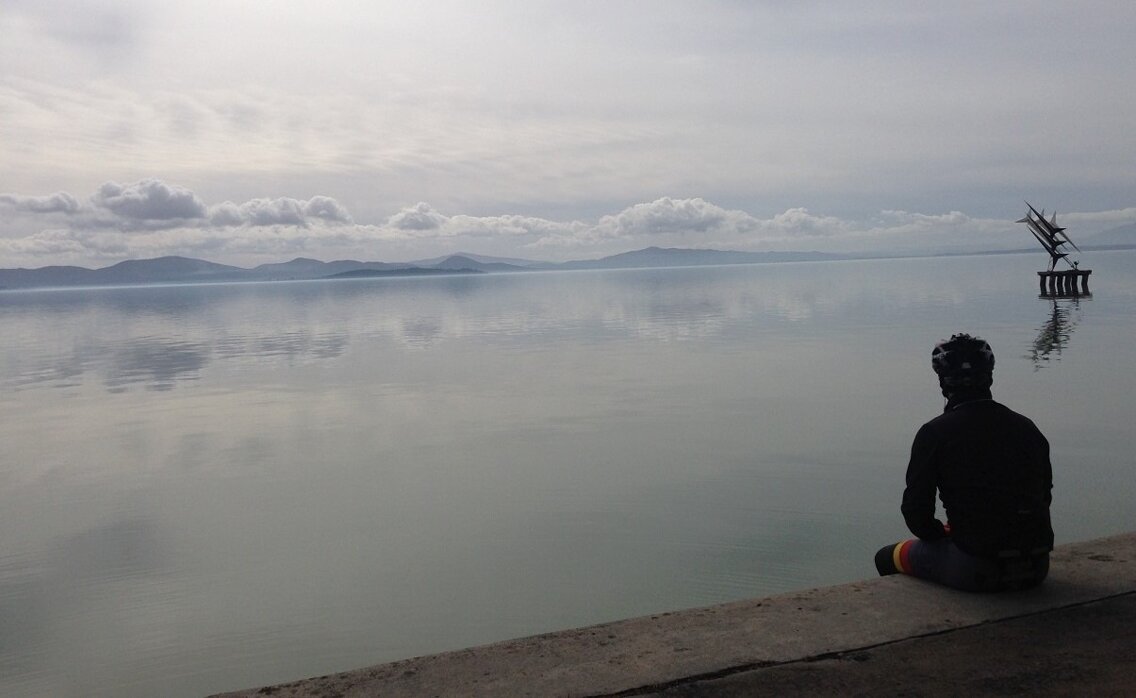 il muretto a secco del lungo lago che si affaccia sul lago trasimeno, la sagoma di spalle del ciclista che osserva il lago su cui si riflettono le nuvole ed una scultura posta al centro del lago, qualche ora prima del tramonto
