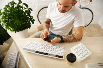 young-tattooed-freelancer-blank-white-t-shirt-uses-his-mobile-phone-near-his-computer-home-front-brick-wall-parked-vintage-bike-summer-time