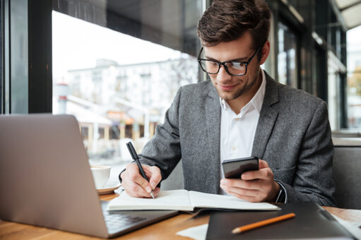 smiling-businessman-eyeglasses-sitting-by-table-cafe-with-laptop-computer-while-using-smartphone-writing-something.jpeg