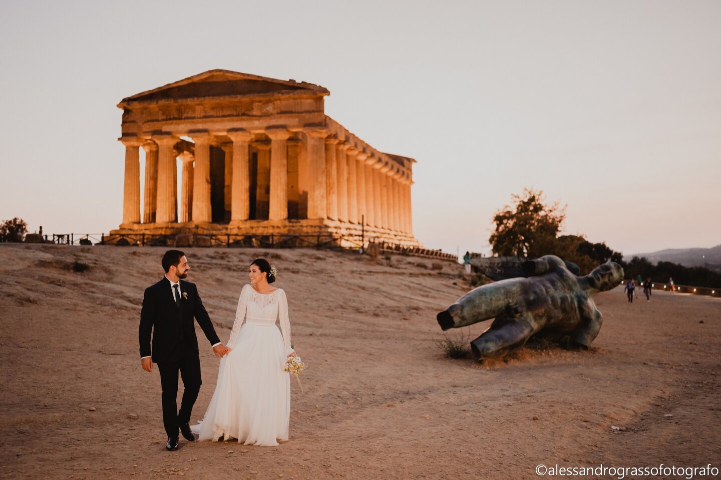 Bride and groom walking near ancient temple in Sicily at sunset.