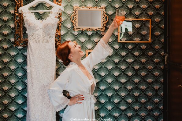Bride celebrating in robe next to her wedding dress, holding a glass of wine.