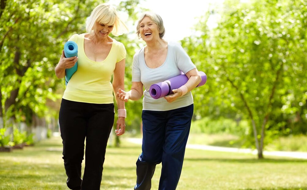Due donne anziane camminano allegre con tappetini yoga in un parco Due donne anziane camminano allegre con tappetini yoga in un parco