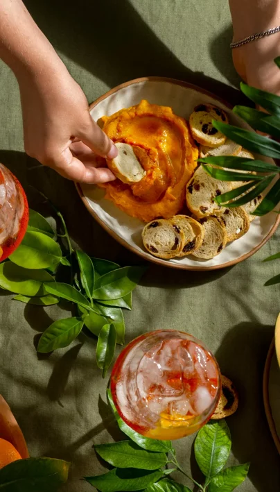 Aperitivo con hummus di peperoni e crostini su tavola di stoffa verde.