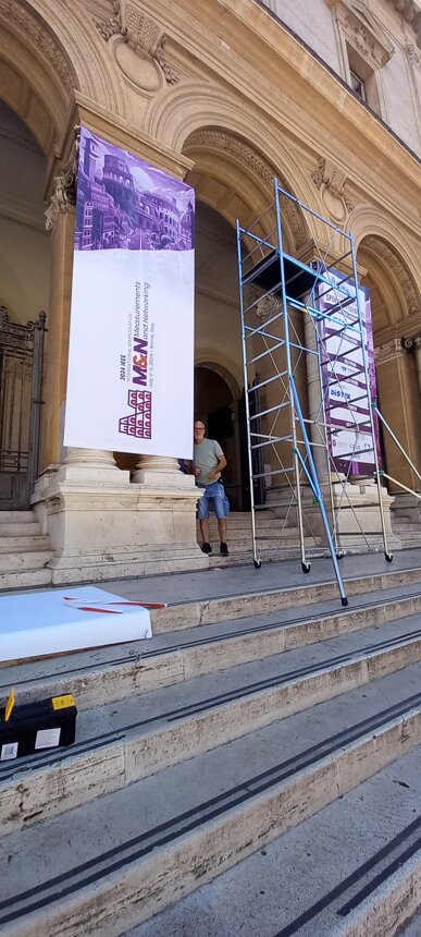 Allestimento di uno striscione per conferenza IEEE MCNN con impalcatura e persona su scalini Allestimento di uno striscione per conferenza IEEE MCNN con impalcatura e persona su scalini