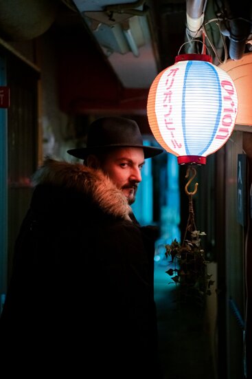 Murdah with fedora hat seen in profile next to a Japanese paper lantern at night murdah srvc photo