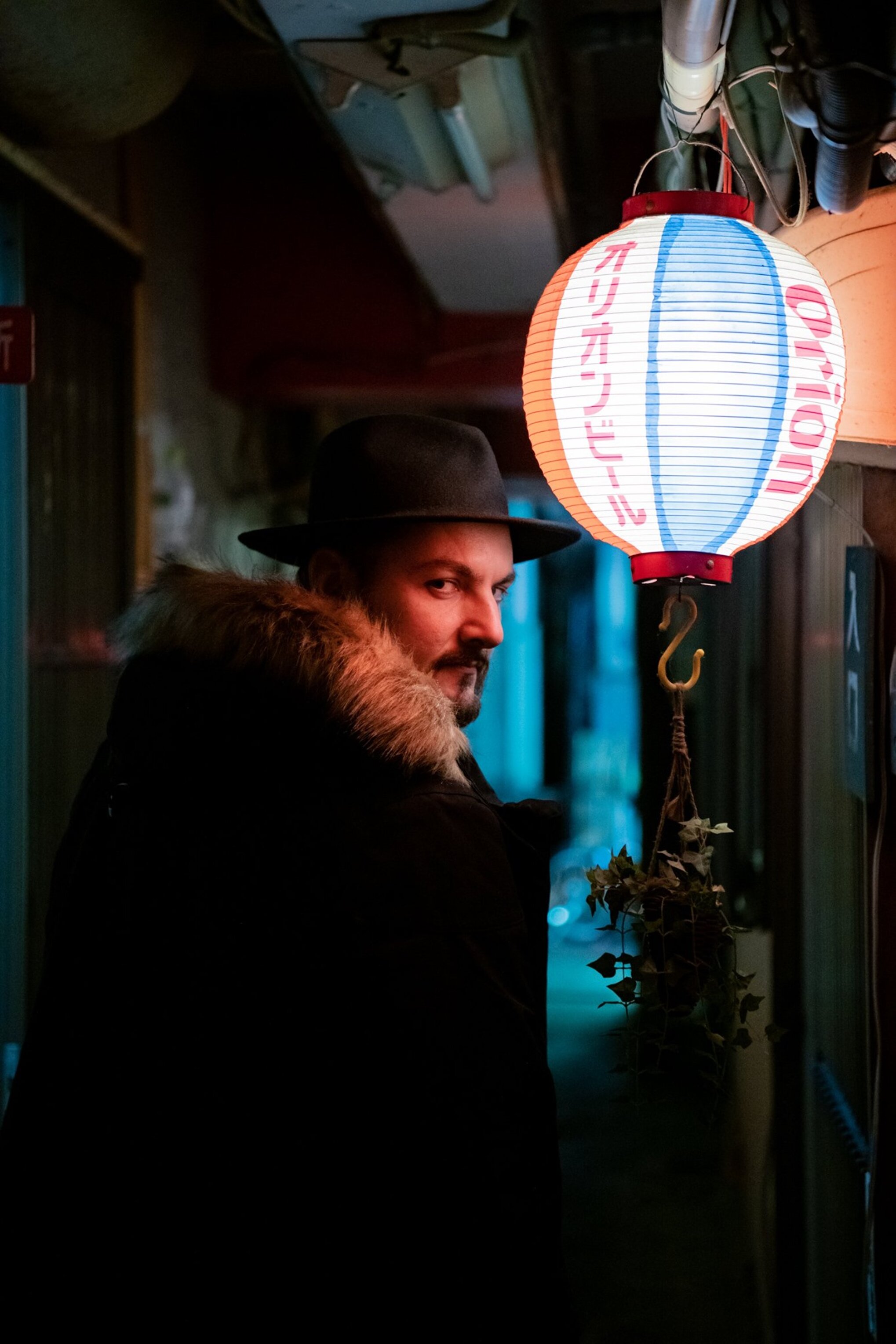 Murdah with fedora hat seen in profile next to a Japanese paper lantern at night murdah srvc photo