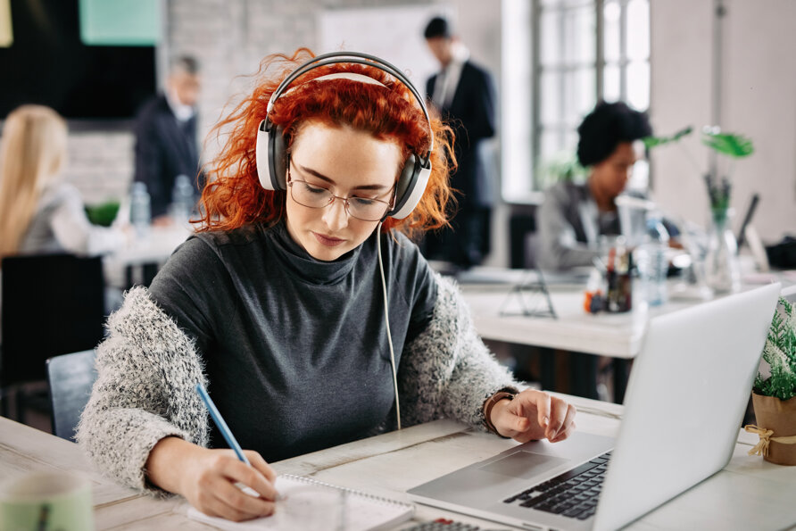 redhead-businesswoman-using-laptop-writing-notes-her-notepad-while-listening-music-headphones-work.jpeg