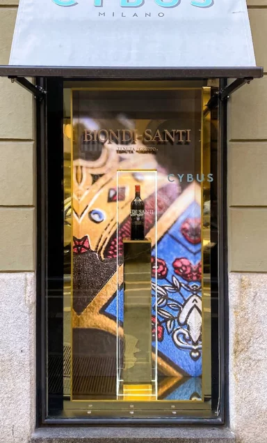 Ingegnerizzazione e allestimento della vetrina luxury Biondi Santi presso Cybus Milano: struttura in specchio oro e plexiglass retroilluminato nel Quadrilatero della Moda. Sviluppo strutturale per una vetrina nel distretto del lusso milanese. L'intervento inizia con il rivestimento integrale del vano preesistente in tessuto nero logato. La produzione in-house si focalizza su un box espositivo custom in legno, rivestito in specchio oro e incastonato al millimetro. Il podio per il Brunello fonde una colonna centrale specchiata, inglobata in una struttura in plexiglass con profili incisi e illuminati al laser. Il setup tecnico si chiude con un fondale grafico retroilluminato.