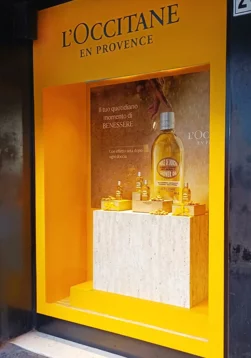 Produzione in-house di prop scenografici e visual merchandising esecutivo per il roll-out vetrine L'Occitane in Coin Roma, Sviluppo strutturale per display product-centric. Ingegnerizzazione e manifattura in-house di un box espositivo multi-livello. Finiture custom in finto travertino e laccature gialle per massimizzare il contrasto visivo del prodotto. 